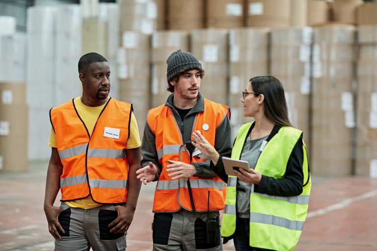 Waist-up view of Caucasian and Black male workers in 20s and 40s wearing reflective vests and walking with supervisor discussing storage management.