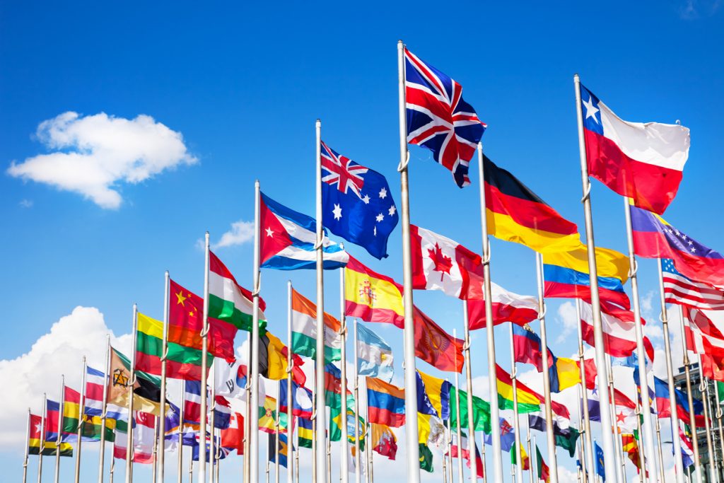 Group of flags of many different nations against blue sky and in front of a convention centre.