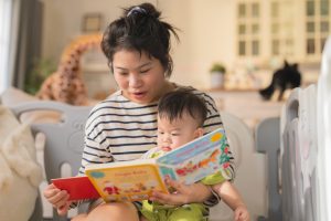 Young loving mother reading bedtime storybook to adorable baby Asian baby boy sitting on Grandmother's lap and reading a picture book family lifestyle at home