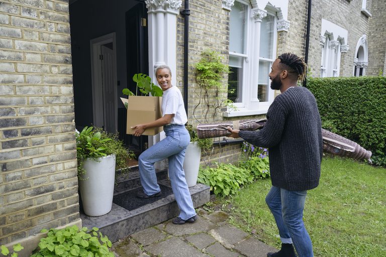 Full length view of man and woman in casual clothing smiling at each other as they carry cardboard box and rug up front steps on moving day.